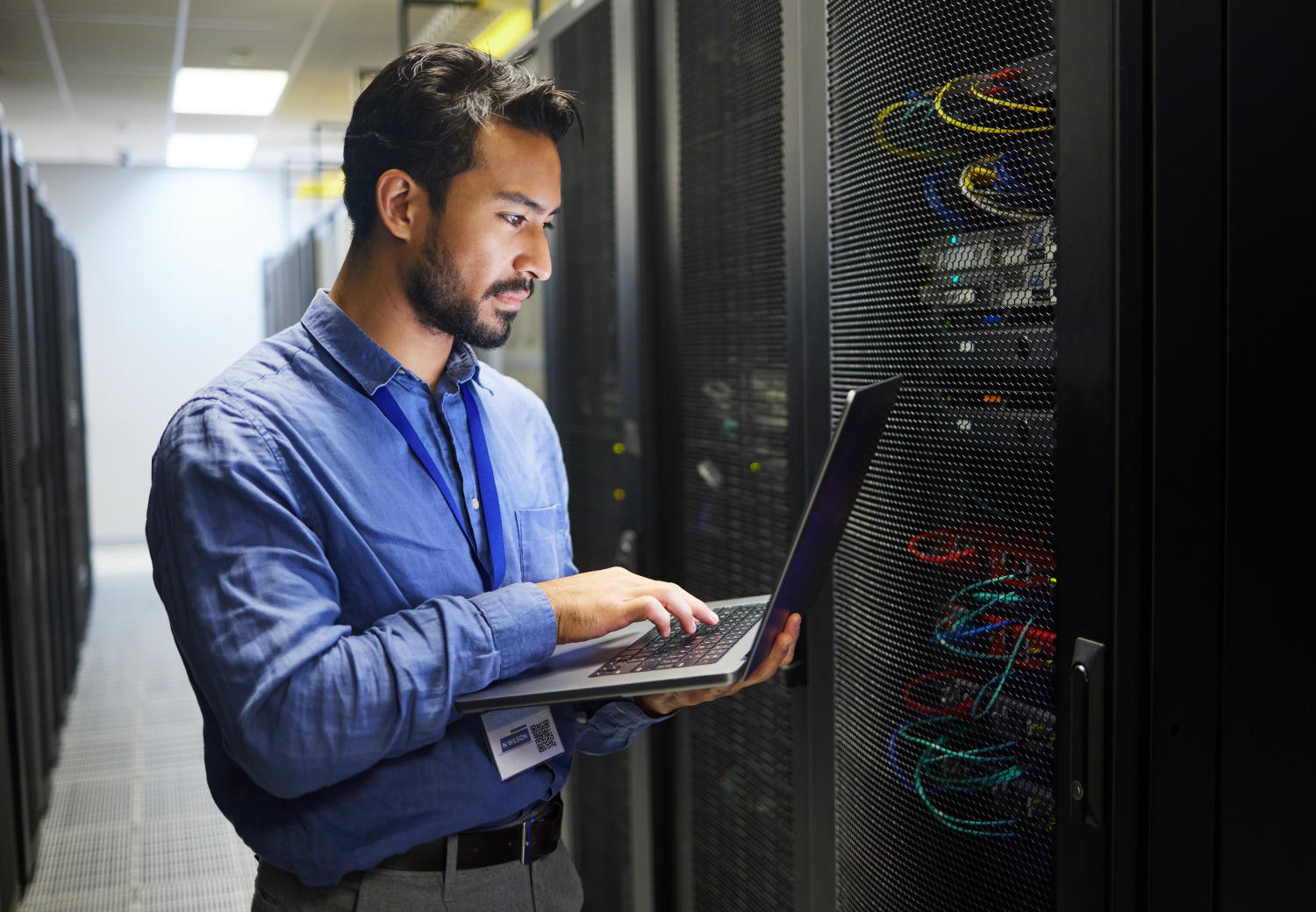 Mann steht vor Serverschrank mit Laptop in der Hand